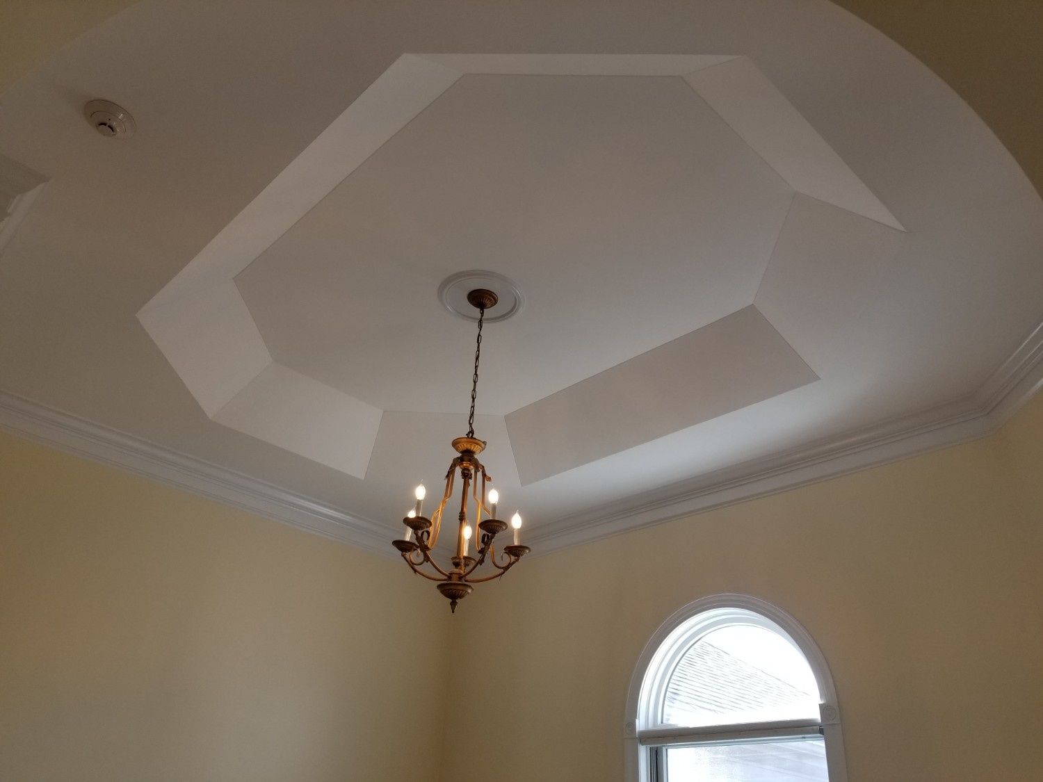 A chandelier hangs from a geometric, recessed tray ceiling in a room with light yellow walls and an arched window.