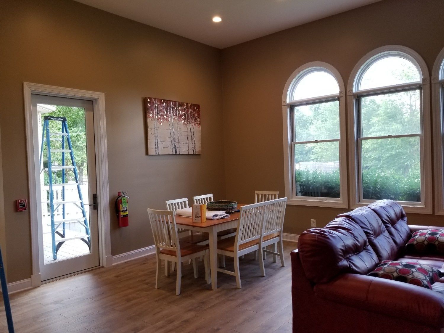 A room with a wooden dining table, white chairs, a brown leather sofa, and three arched windows overlooking greenery.