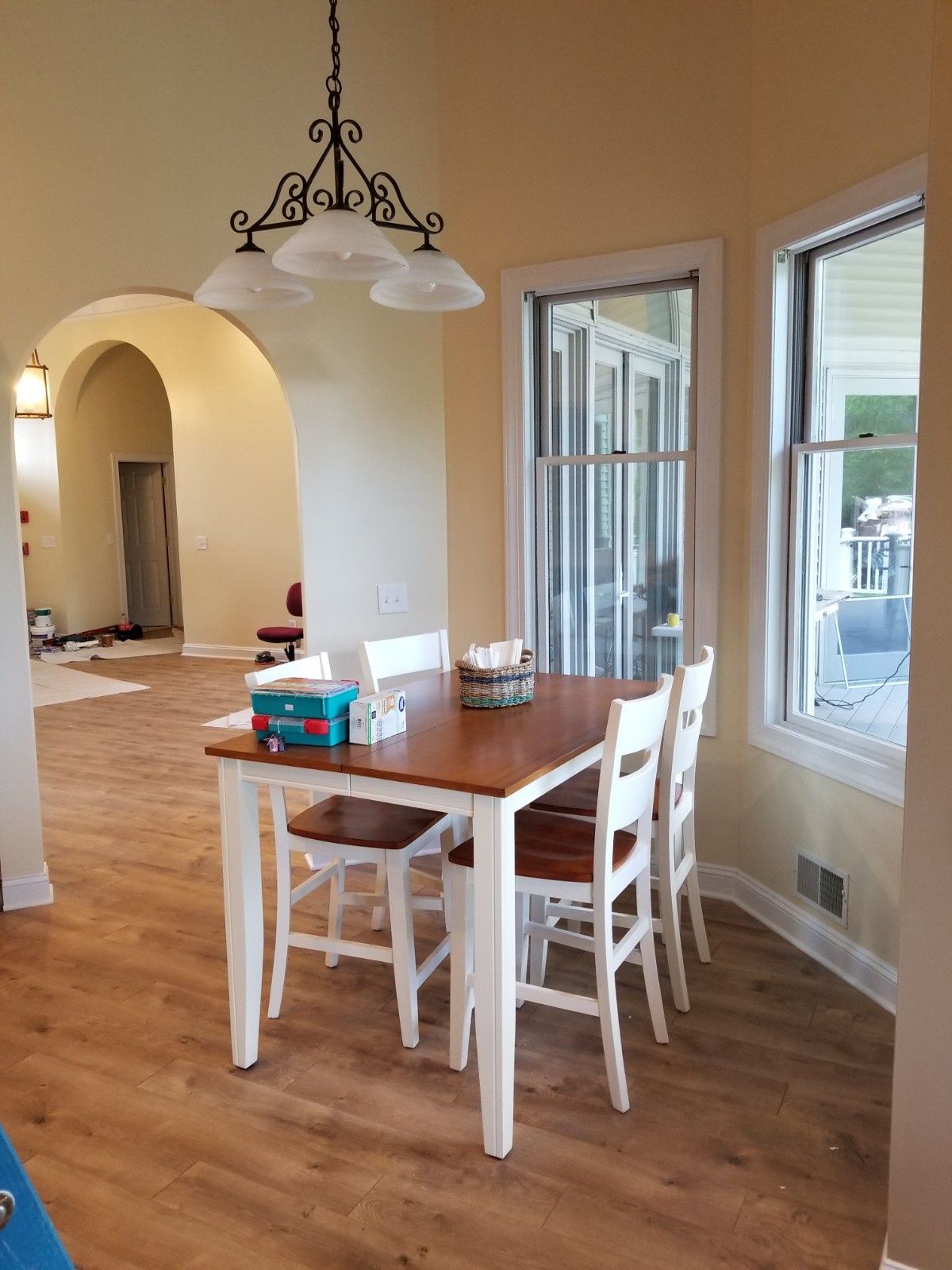 A kitchen nook with a wooden dining table and white chairs under a decorative chandelier, next to windows with outdoor views.