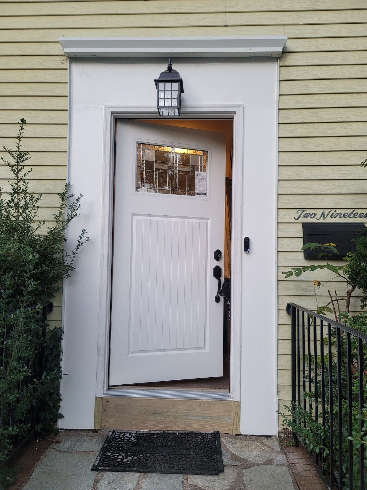 A white wooden front door slightly ajar on a light yellow clapboard house with a small black porch light above.