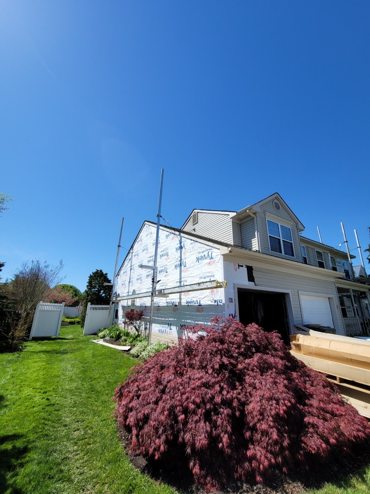 A house under construction with scaffolding against a white-wrapped exterior wall, next to a large red-leafed bush.
