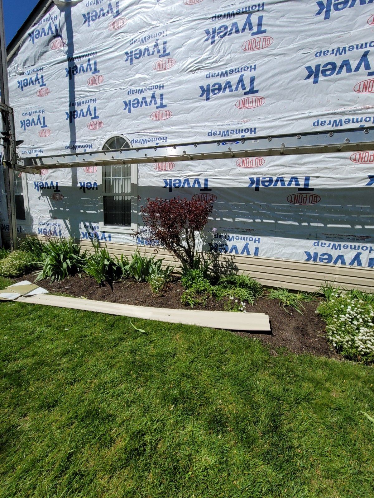 A house exterior under construction, featuring Tyvek house wrap, a window, scaffolding, and a landscaped garden bed.