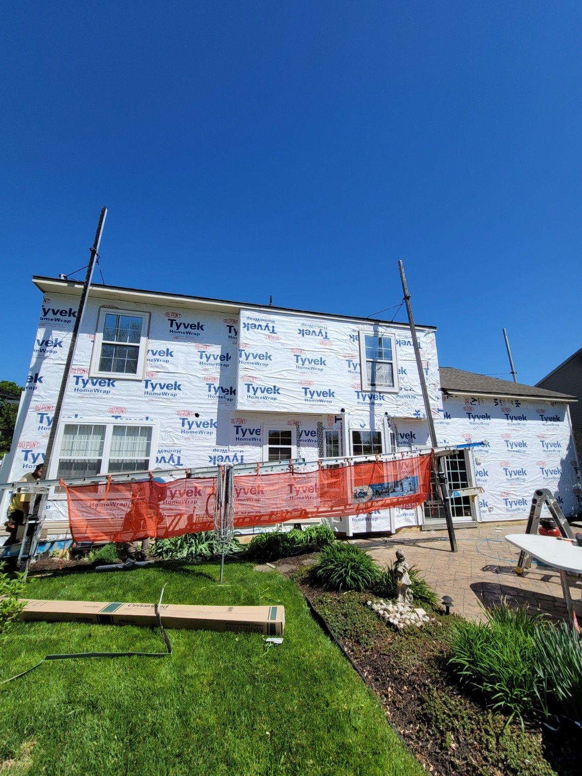 The back of a two-story home undergoing exterior renovation, covered in Tyvek house wrap with scaffolding and orange mesh.