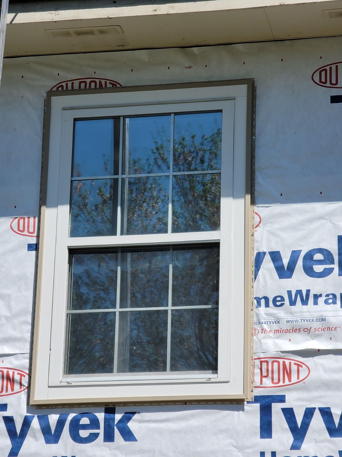 A white-framed window with a grid pattern installed on a building exterior covered in white Tyvek wrap.
