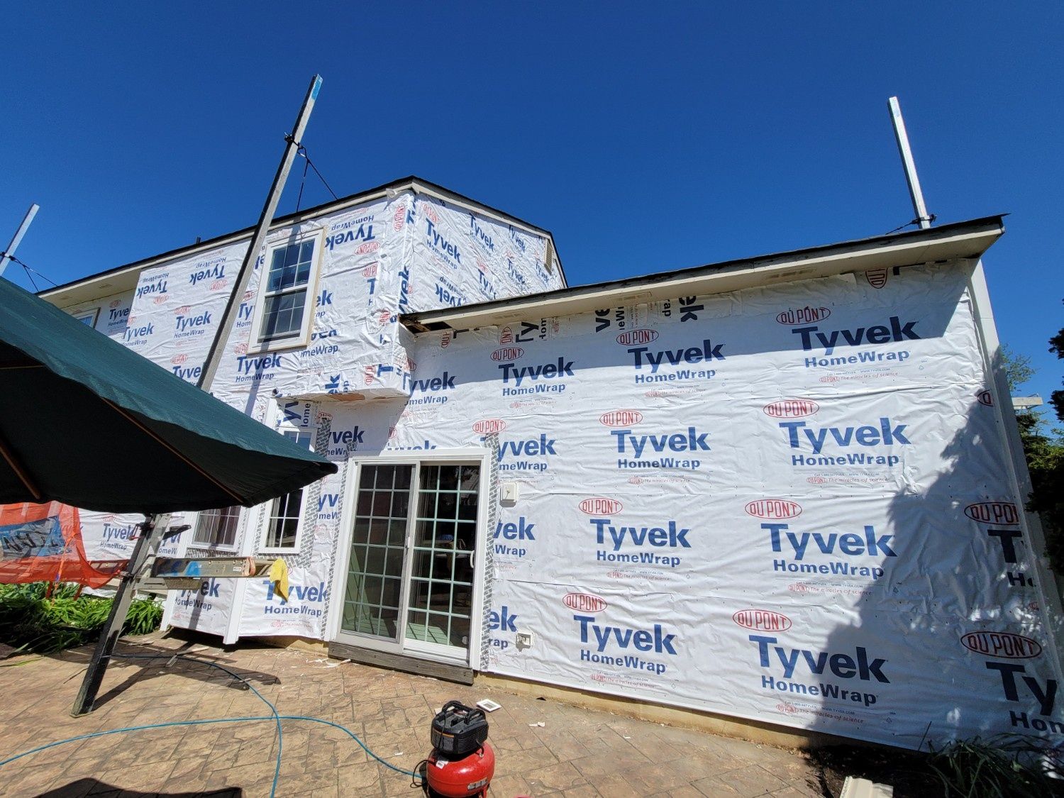 A two-story house exterior under construction, wrapped in white Tyvek weather-resistant barrier, featuring a sliding door.