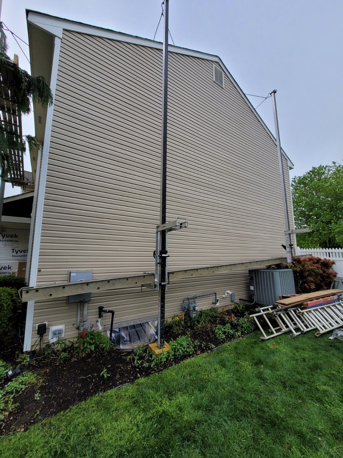 A tan, vinyl-sided house exterior with a metal pole, ladder, and landscaping equipment in the yard.