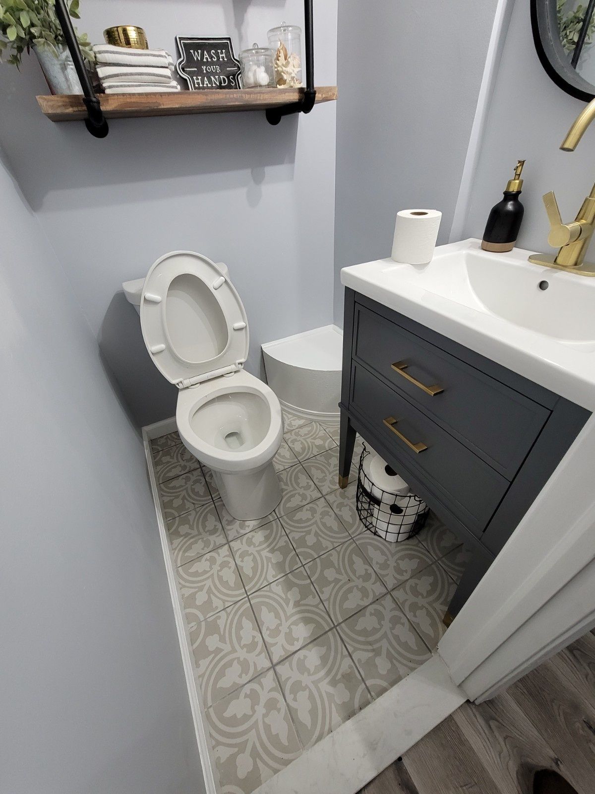 A modern small bathroom featuring a white toilet, gray vanity with gold handles, patterned floor tiles, and wood shelving.