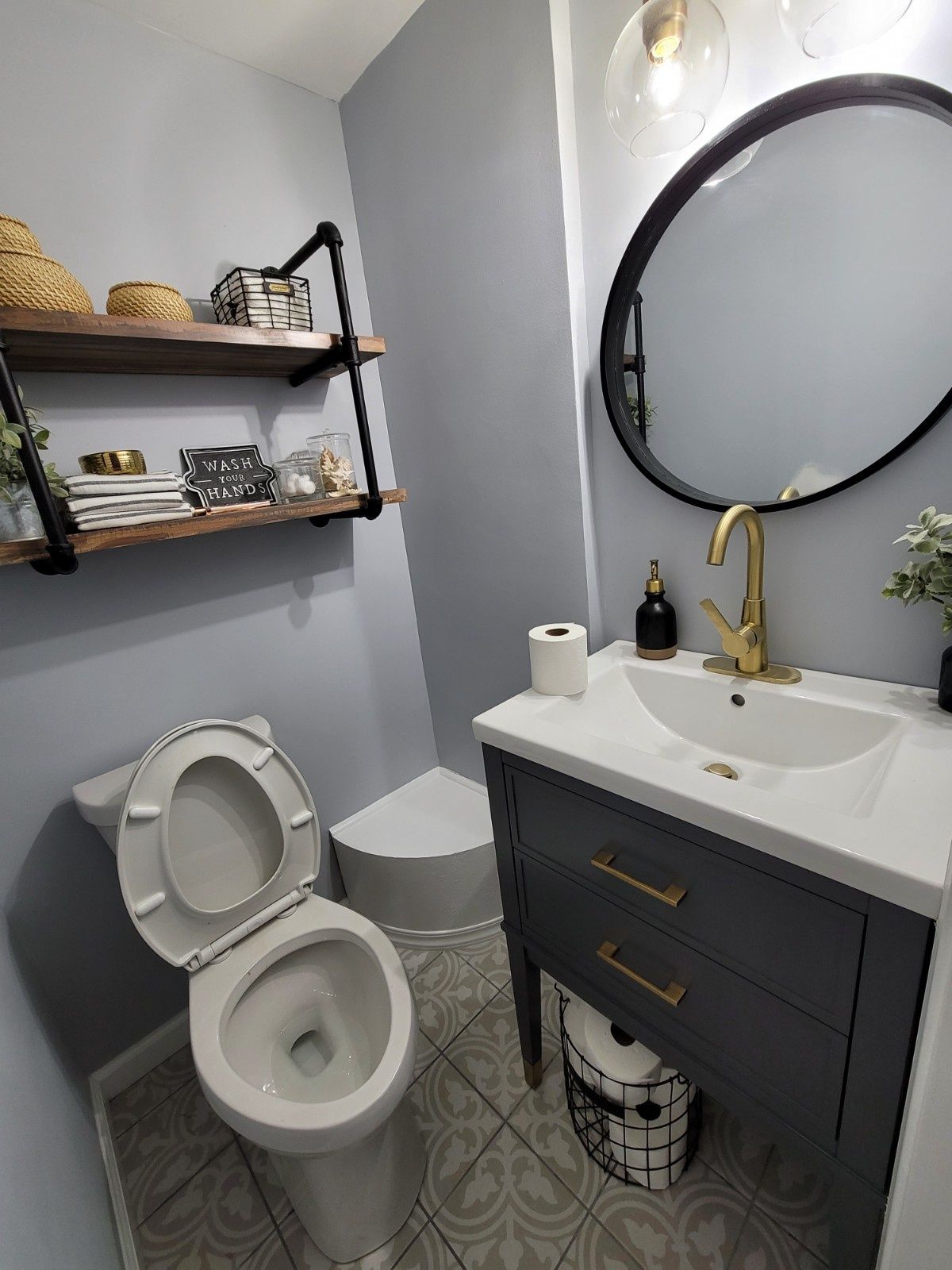 A bathroom featuring a gray vanity with a gold faucet, a round mirror, wooden wall shelves, and a white toilet.