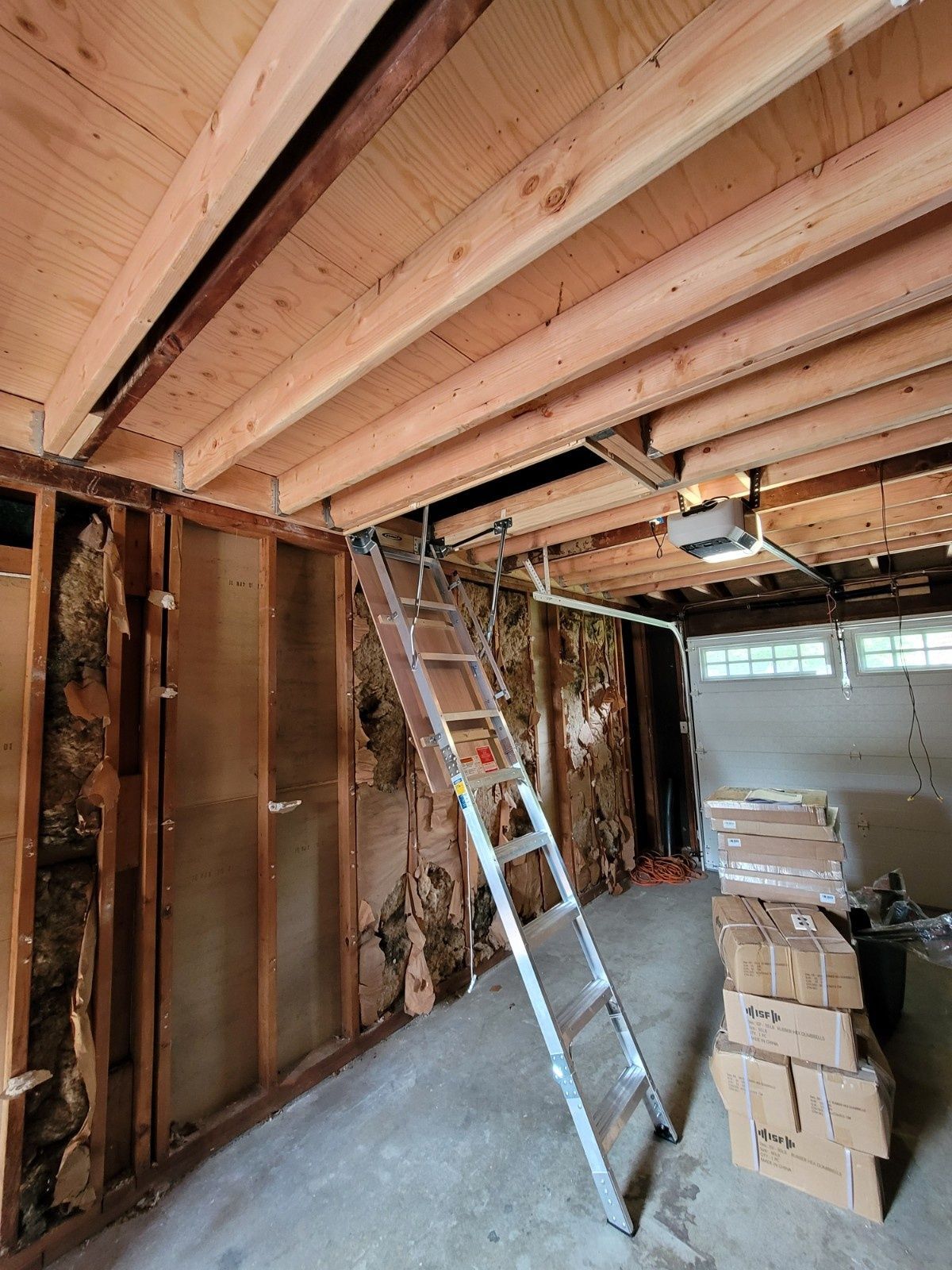 A metal ladder leans against an unfinished garage ceiling, which is exposed to the joists, next to insulation and boxes.