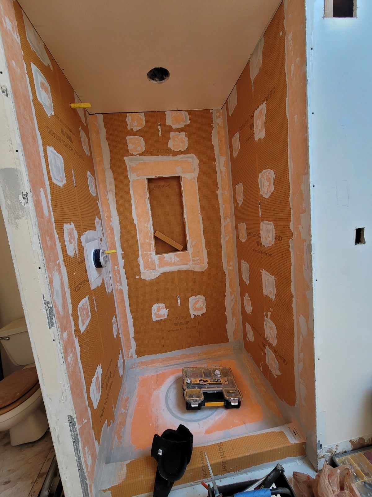 A shower stall under construction, featuring orange waterproof membrane on walls and floor with a central built-in niche.