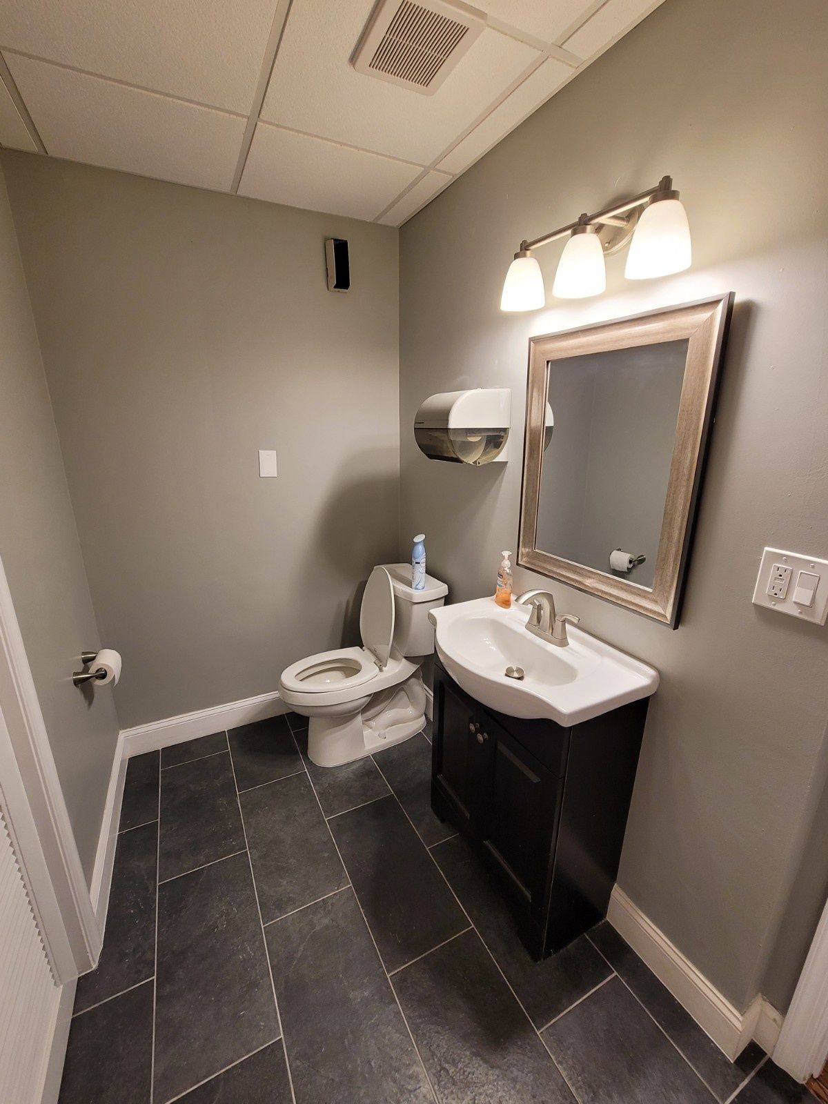 A small, gray-walled bathroom featuring a dark vanity with a white sink, a mirror, and a toilet on a black tiled floor.