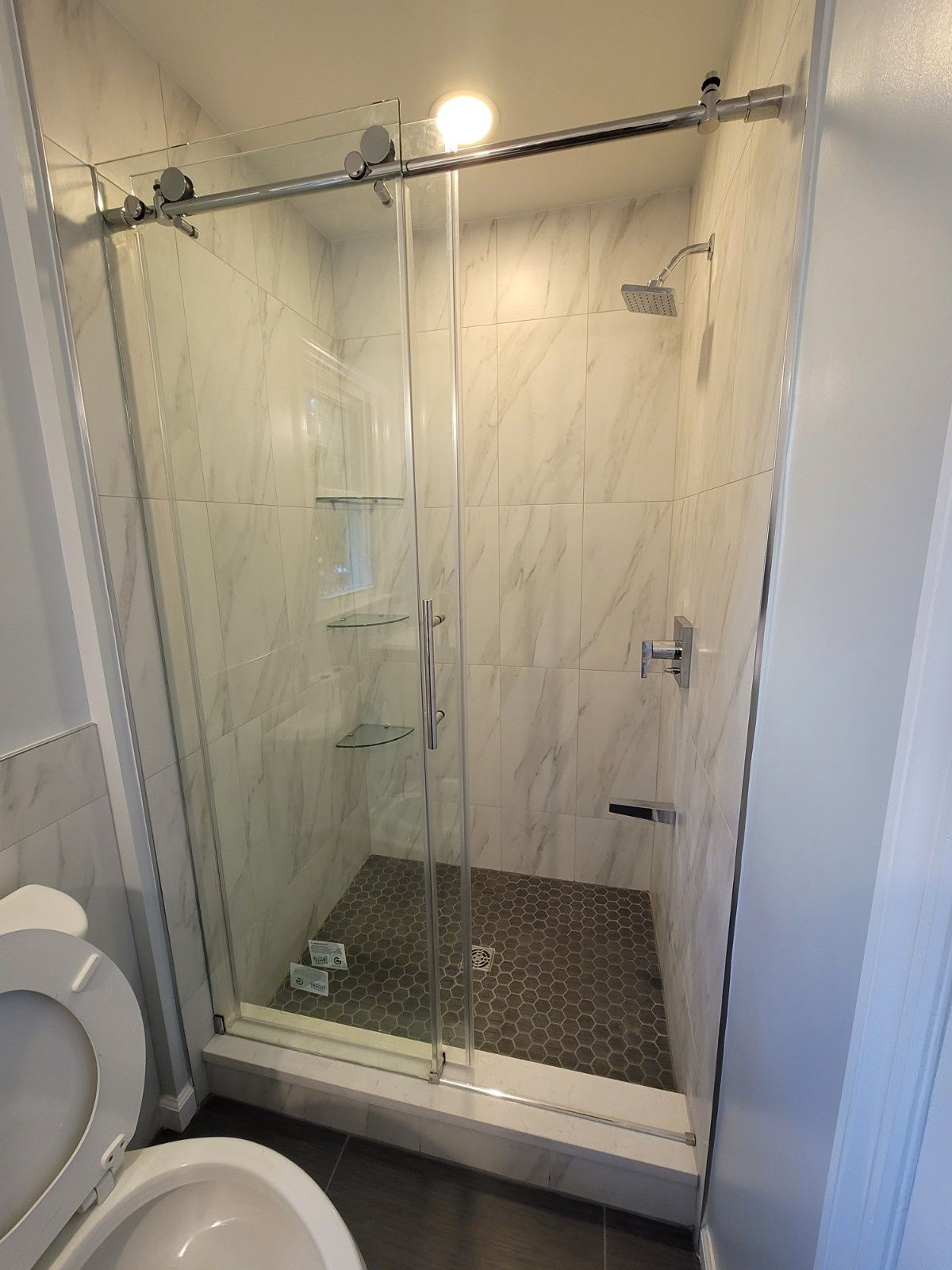 A walk-in shower with a glass sliding door, marble-patterned tile walls, and a dark pebble floor next to a toilet.