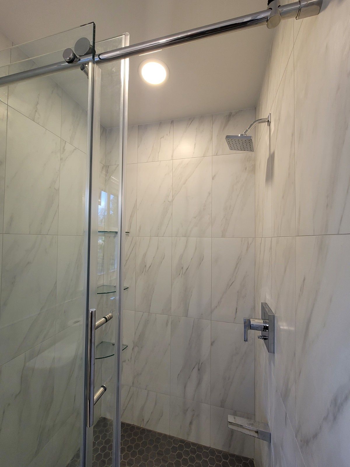 A walk-in shower with white marble-patterned tile walls, a rain showerhead, glass shelves, and a sliding glass door.