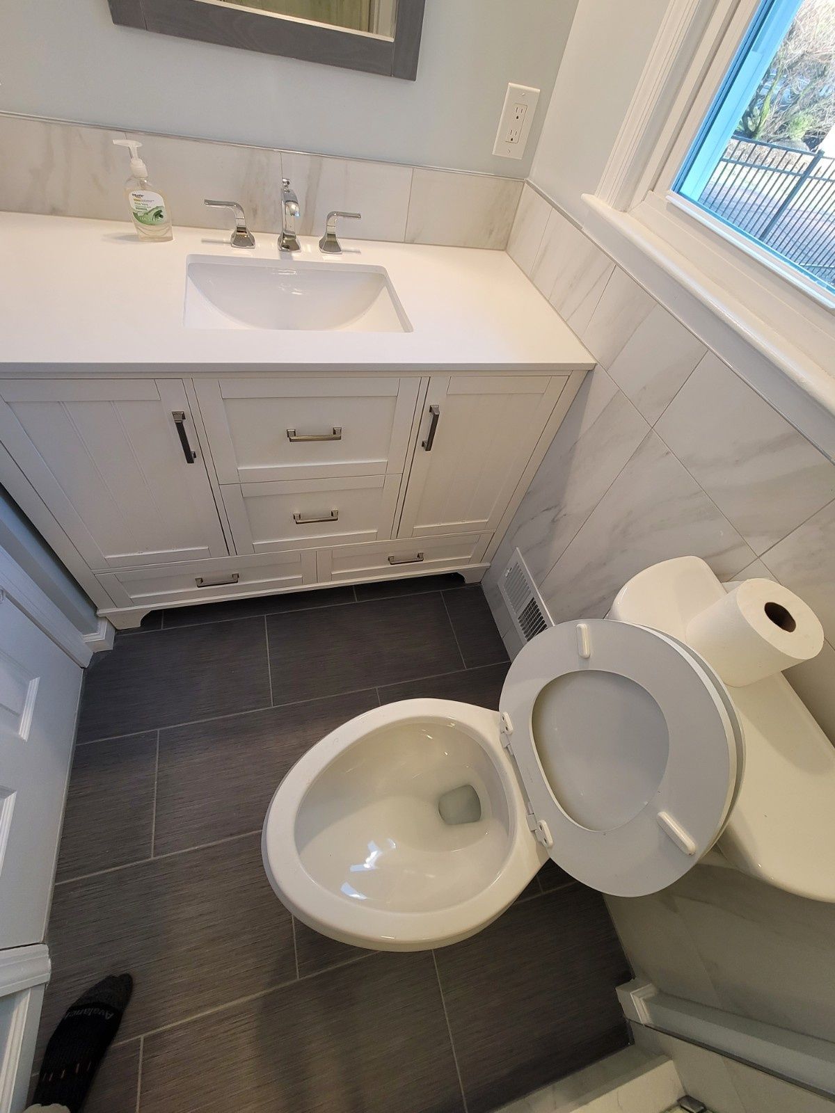 A high-angle view of a bathroom with a white vanity, a white toilet with the lid open, and grey tiled flooring.