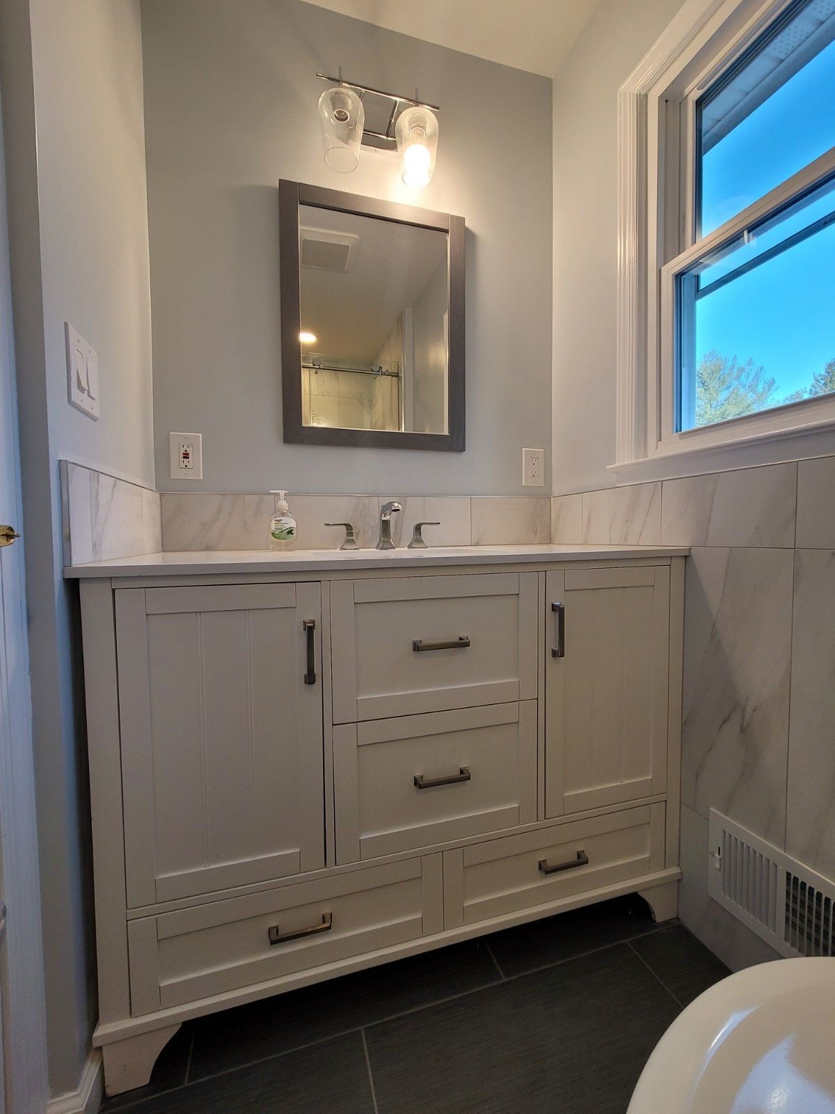 A white bathroom vanity with drawers and a sink beneath a rectangular mirror, set against light gray walls and tiled baseboards.