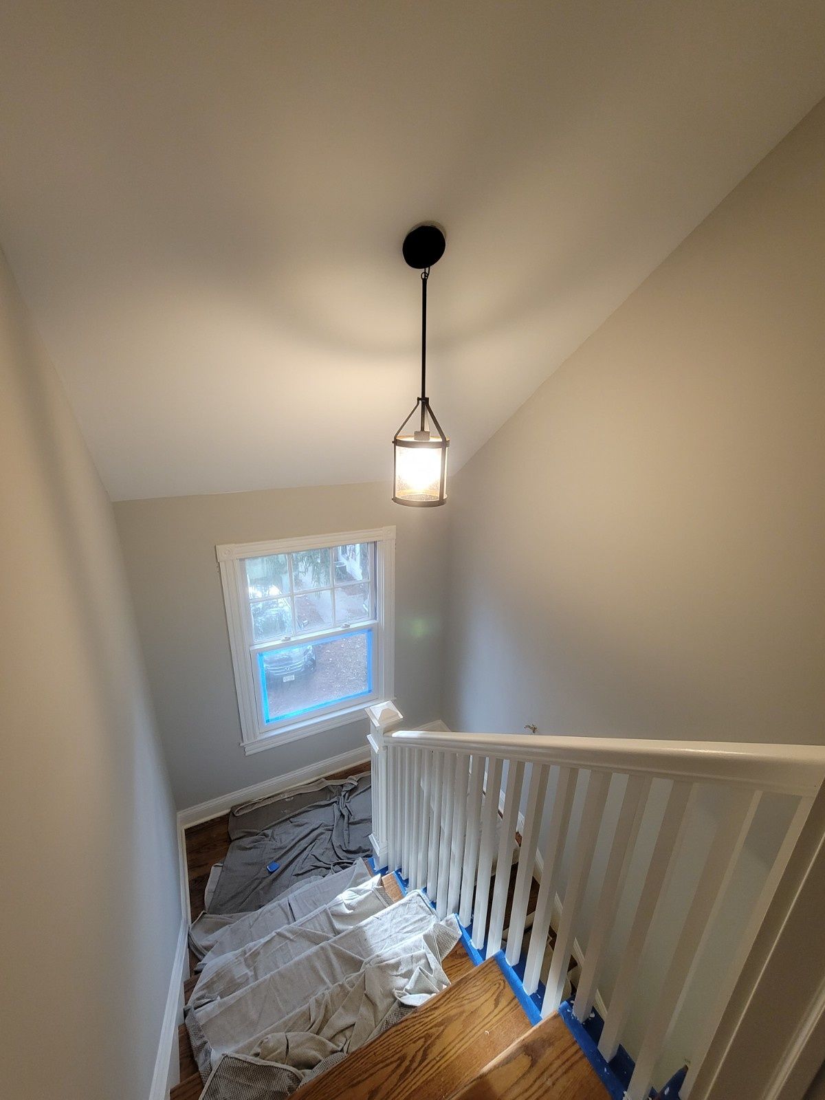 A high-angle view of a staircase with white railings, light gray walls, and a hanging pendant light above a window.