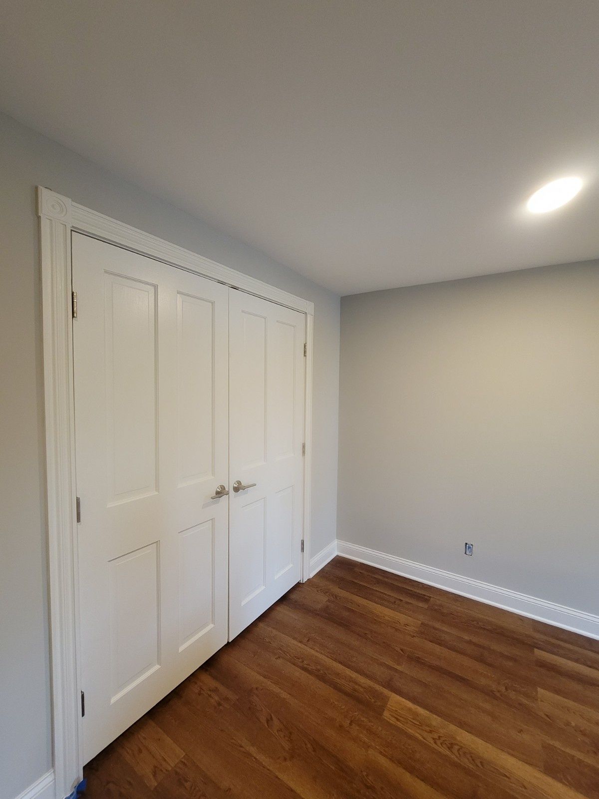 A white double closet door with silver handles stands against a light gray wall in a room with dark wood flooring.