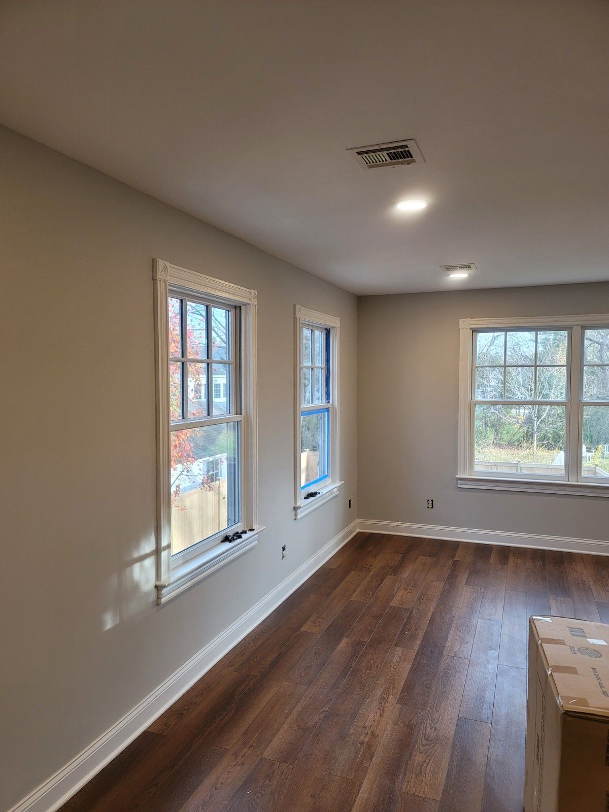 A bright room with light gray walls, white trim, dark wood flooring, and three windows overlooking trees.