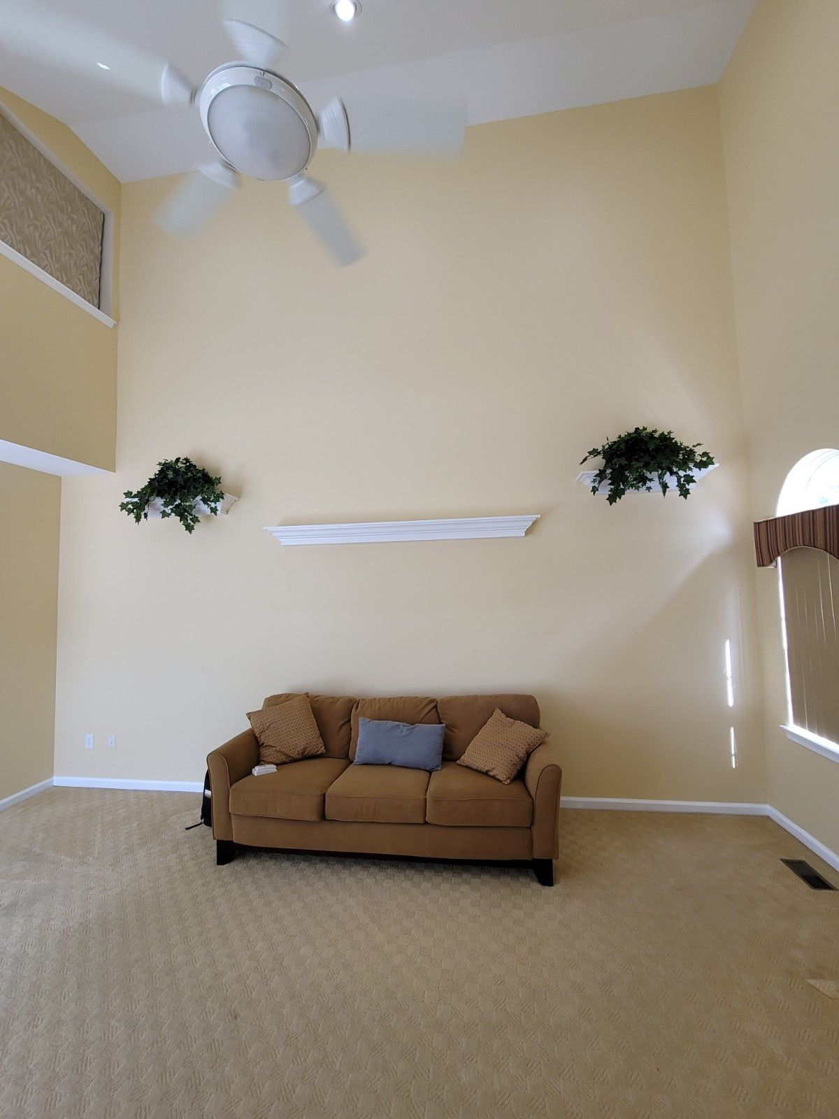 A brown sofa centered below a white mantelpiece on a tall, beige wall in a living room with a ceiling fan.