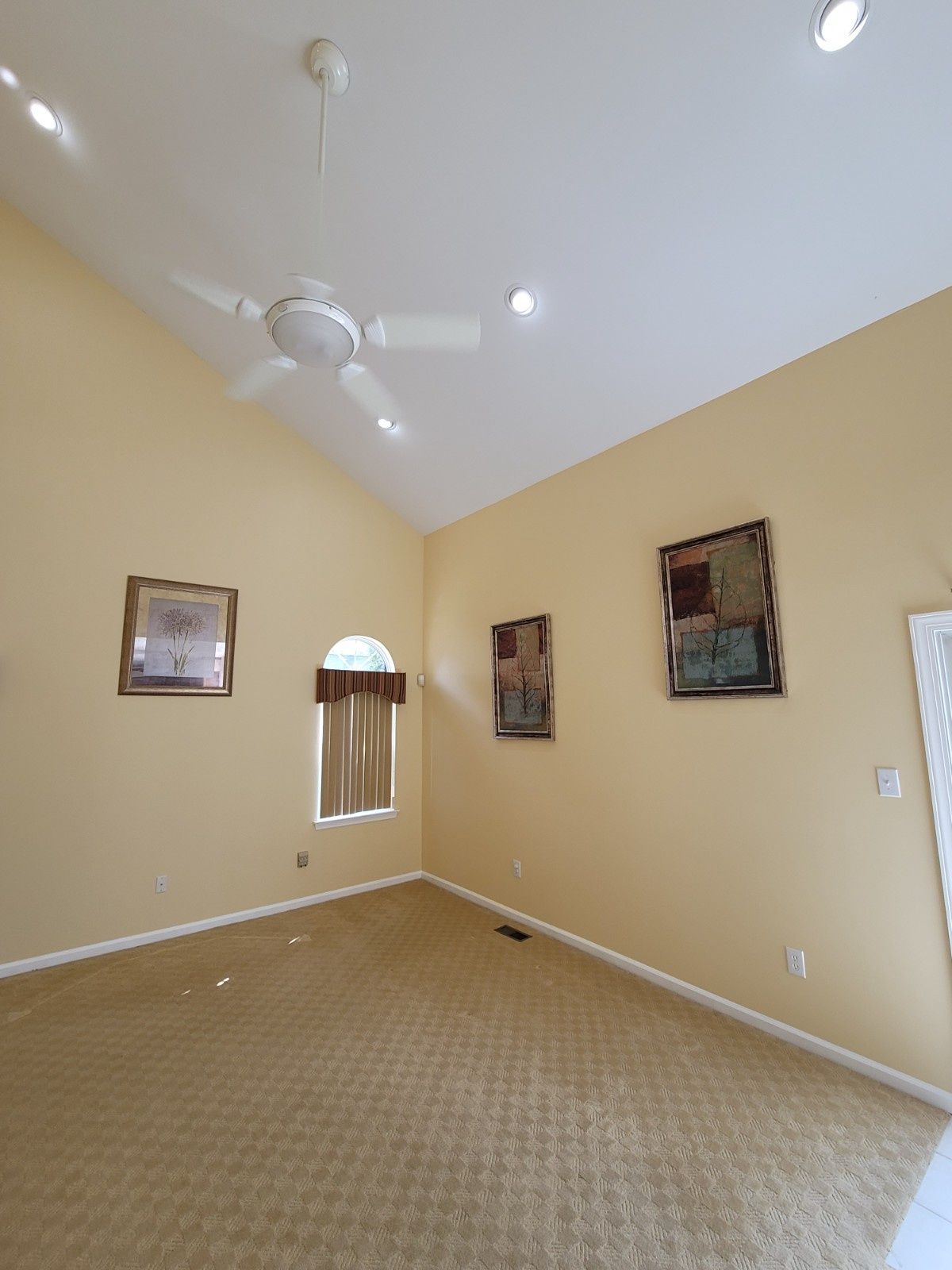 A vacant room with high ceilings, beige walls, carpeted floors, a ceiling fan, a window, and two wall-mounted artworks.