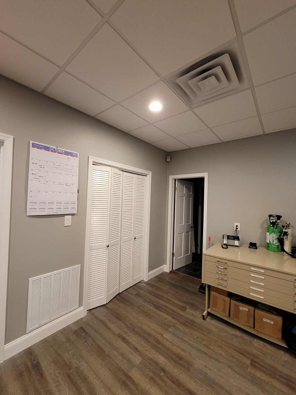 A light gray office interior featuring white folding closet doors, a calendar on the wall, and a cabinet with storage bins.