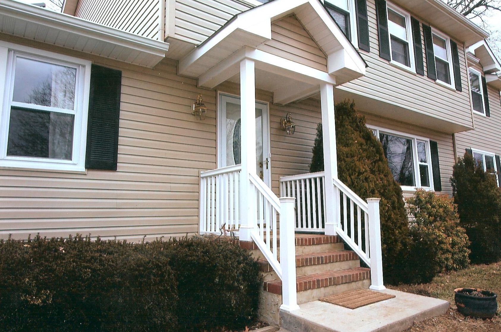 The front entrance of a two-story beige house with white railings, a small covered porch, and landscaping.