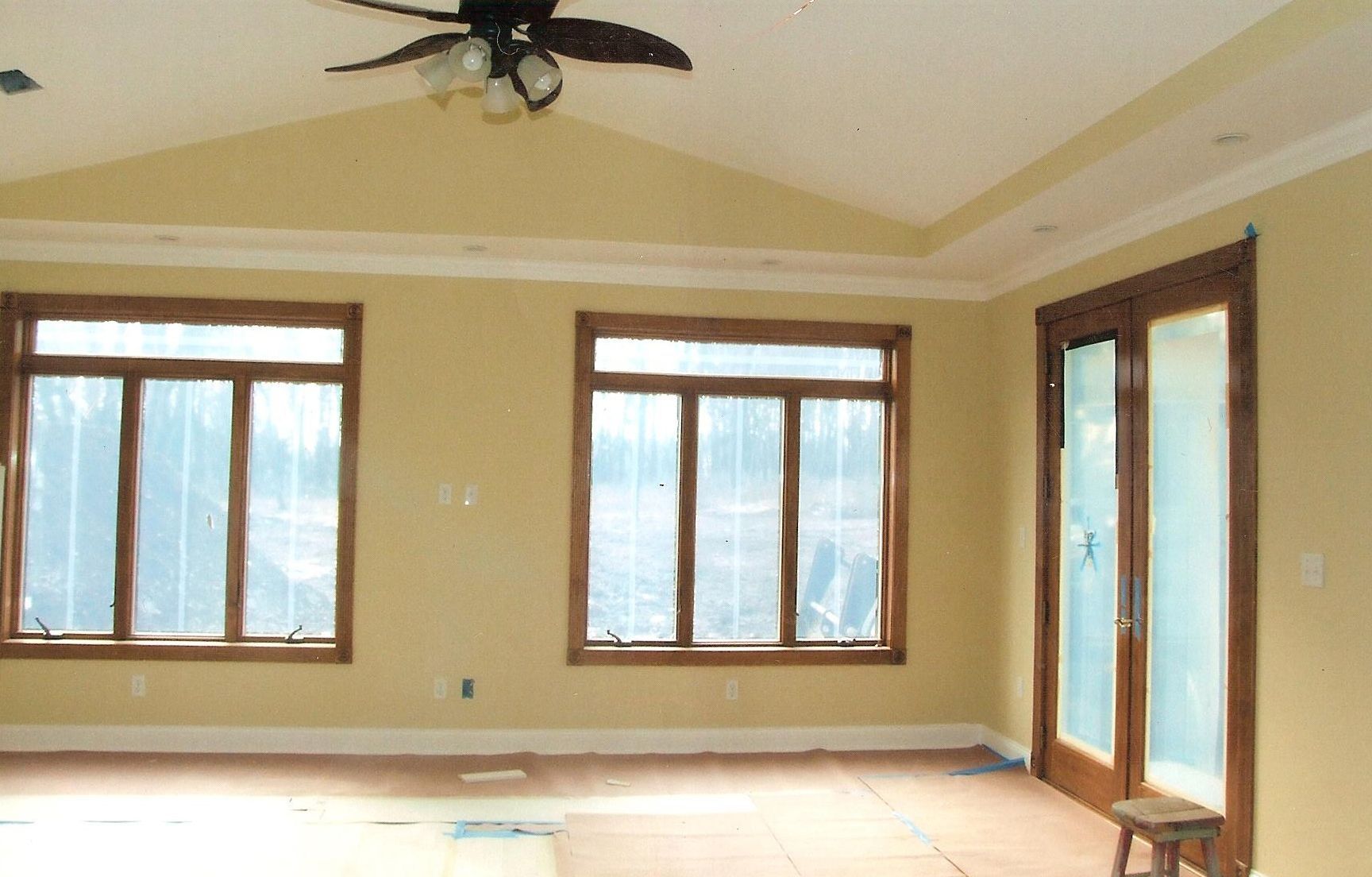 A bright, empty living room with light yellow walls, wood-trimmed windows, a vaulted ceiling, and a ceiling fan.