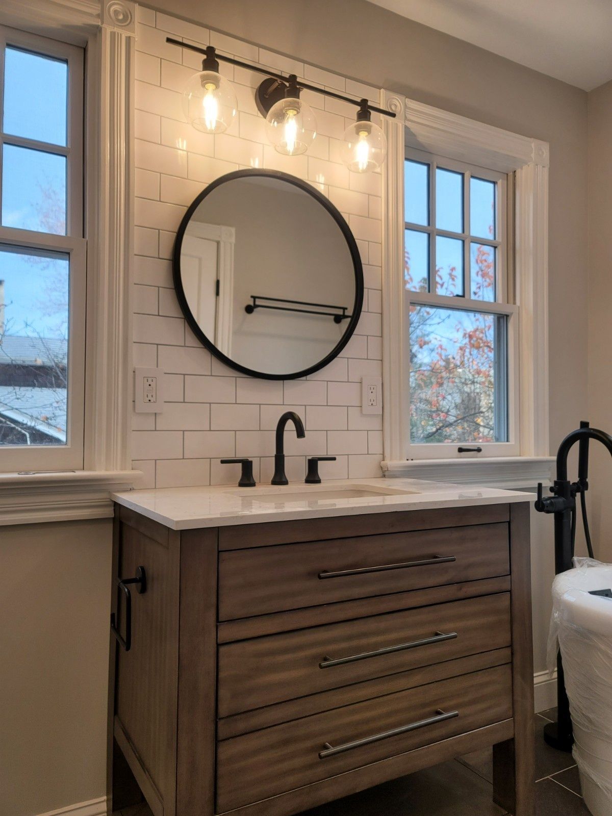 A rustic wooden bathroom vanity with a white countertop, round black mirror, and wall-mounted lighting against subway tiles.