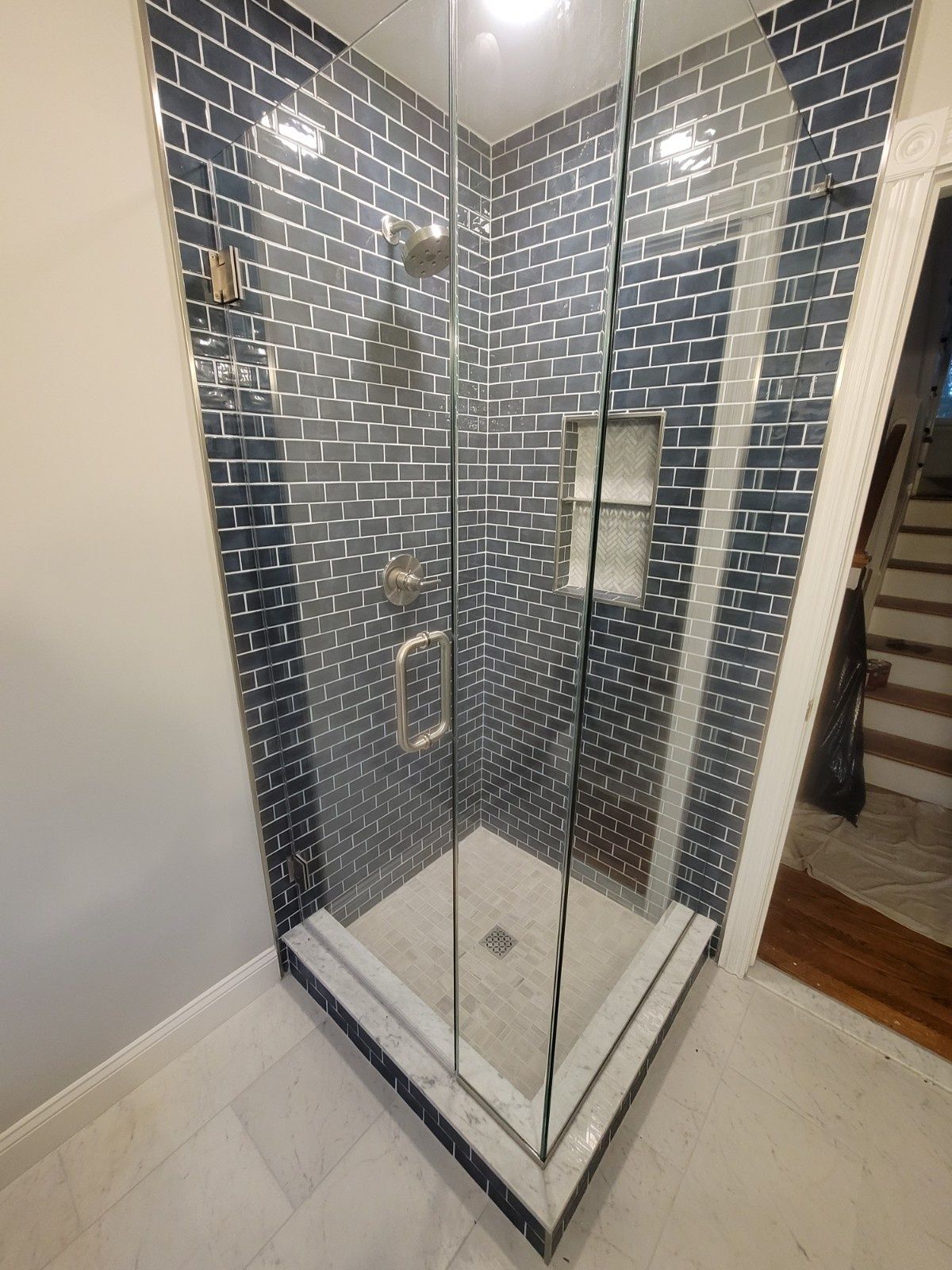 A corner shower with dark blue subway tile walls, a glass enclosure, a built-in shelf, and light-colored stone flooring.