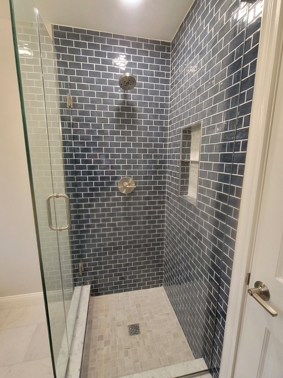 A walk-in shower with dark blue rectangular wall tiles, light floor tiles, a glass door, and a built-in recessed shelf.