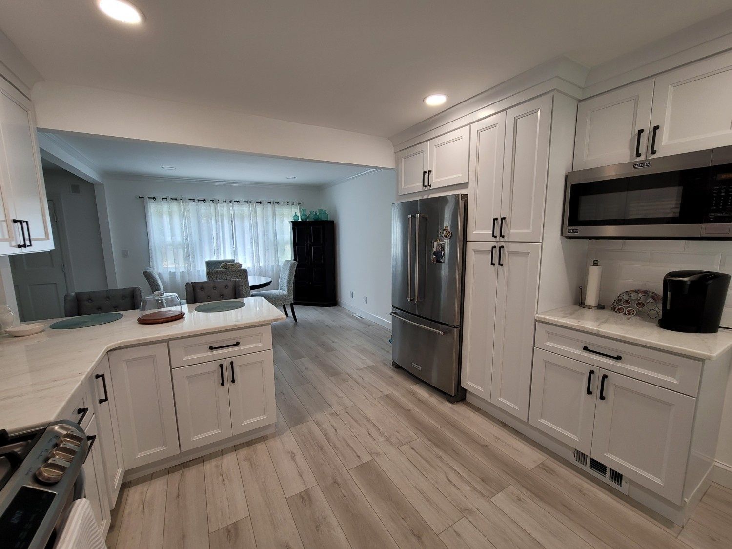 Modern kitchen with white shaker cabinets, stainless steel appliances, and wood-look flooring, leading to a dining area.