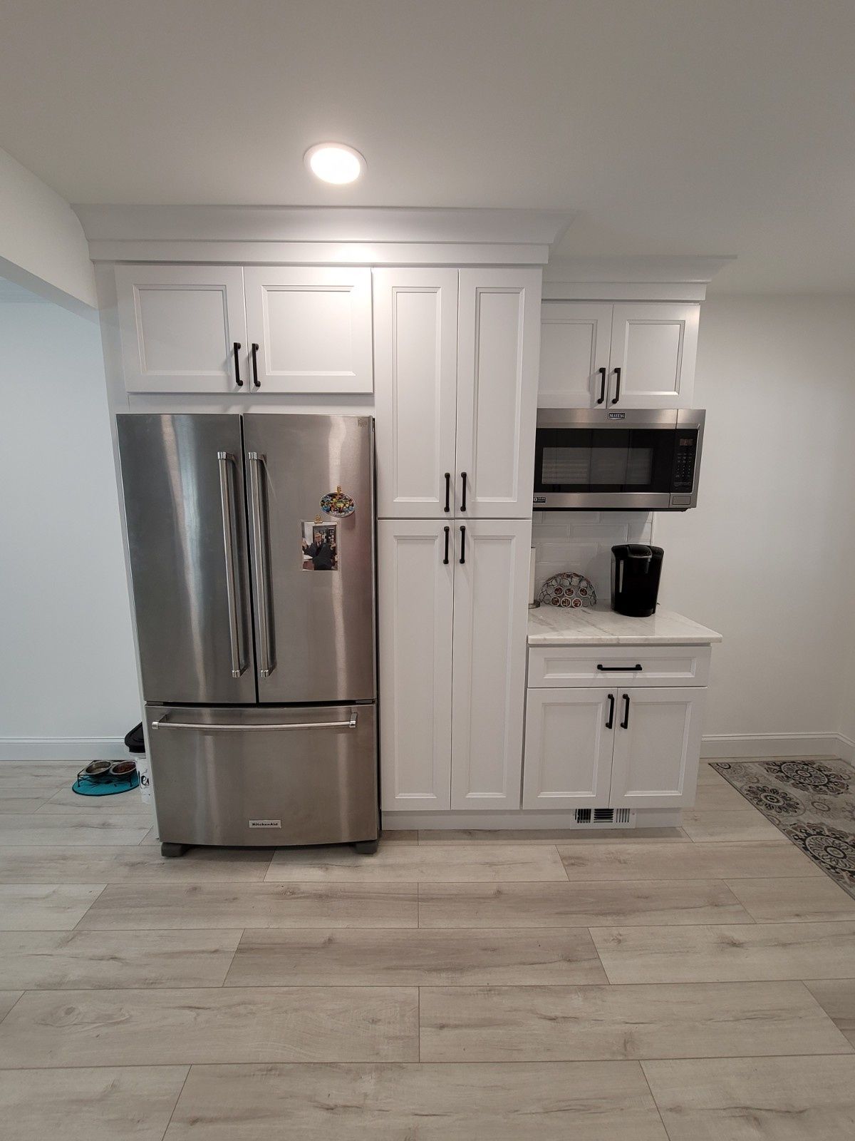 A stainless steel refrigerator sits next to a tall white pantry cabinet and a counter with a microwave and coffee maker.