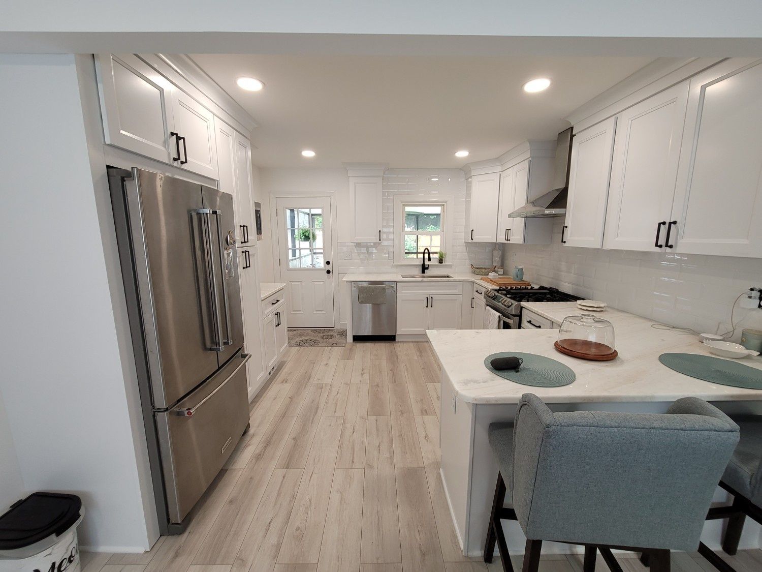 A modern, well-lit kitchen with white cabinets, stainless steel appliances, marble countertops, and light wood flooring.