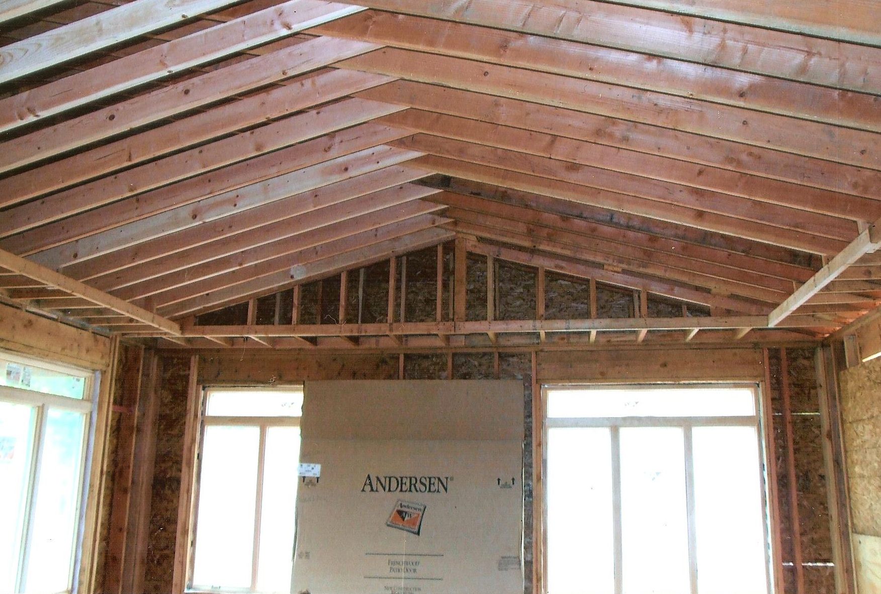 The interior of a house under construction with exposed wooden ceiling rafters, wall studs, and windows.