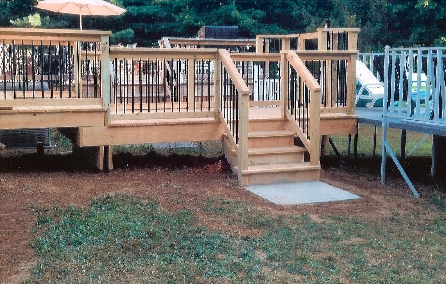 Wooden deck with stairs leading to a backyard, featuring a railing with black metal balusters and a concrete landing.