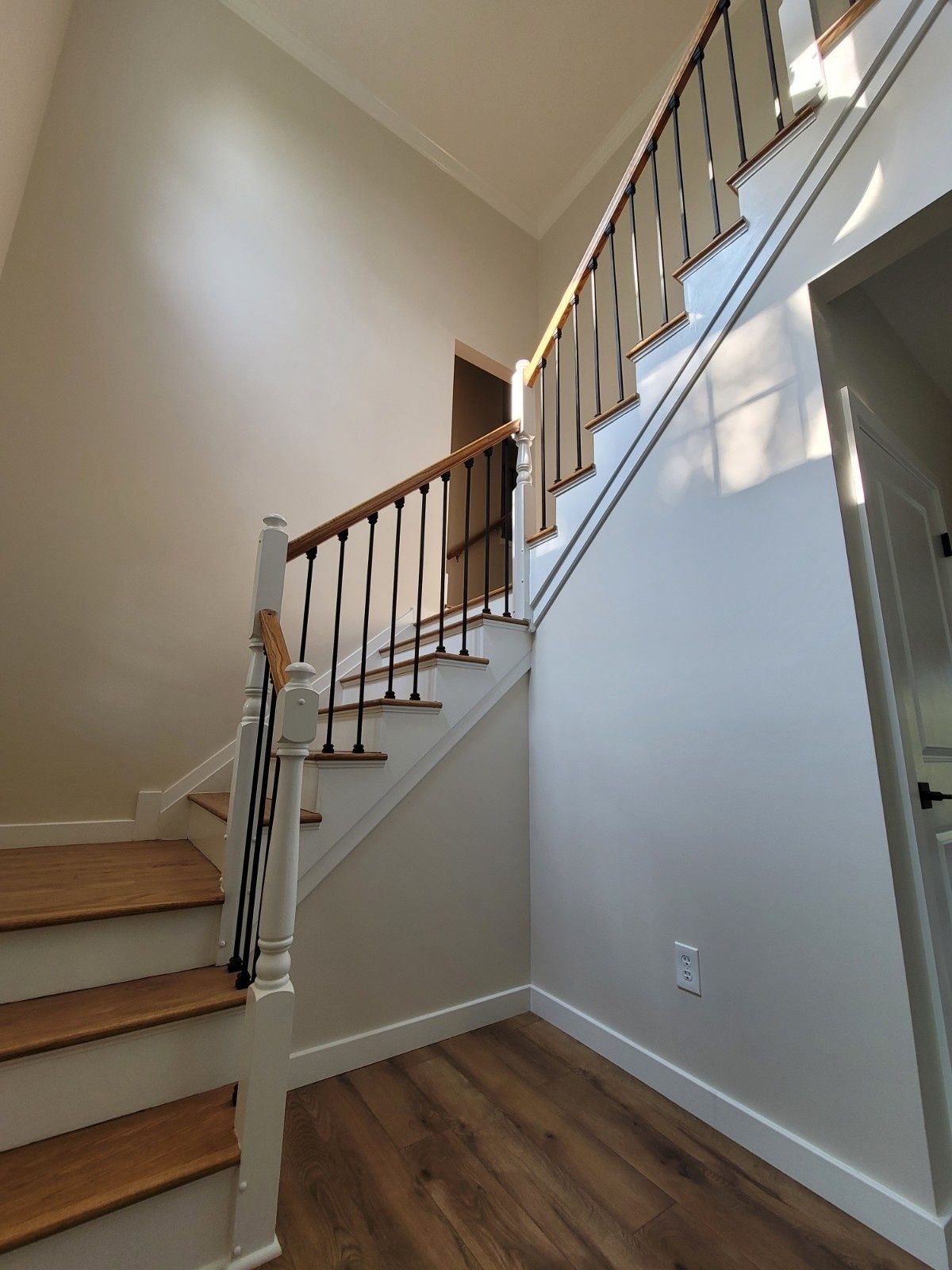 An indoor staircase with wood treads, white risers, and black iron balusters, ascending alongside a tall, light-toned wall.