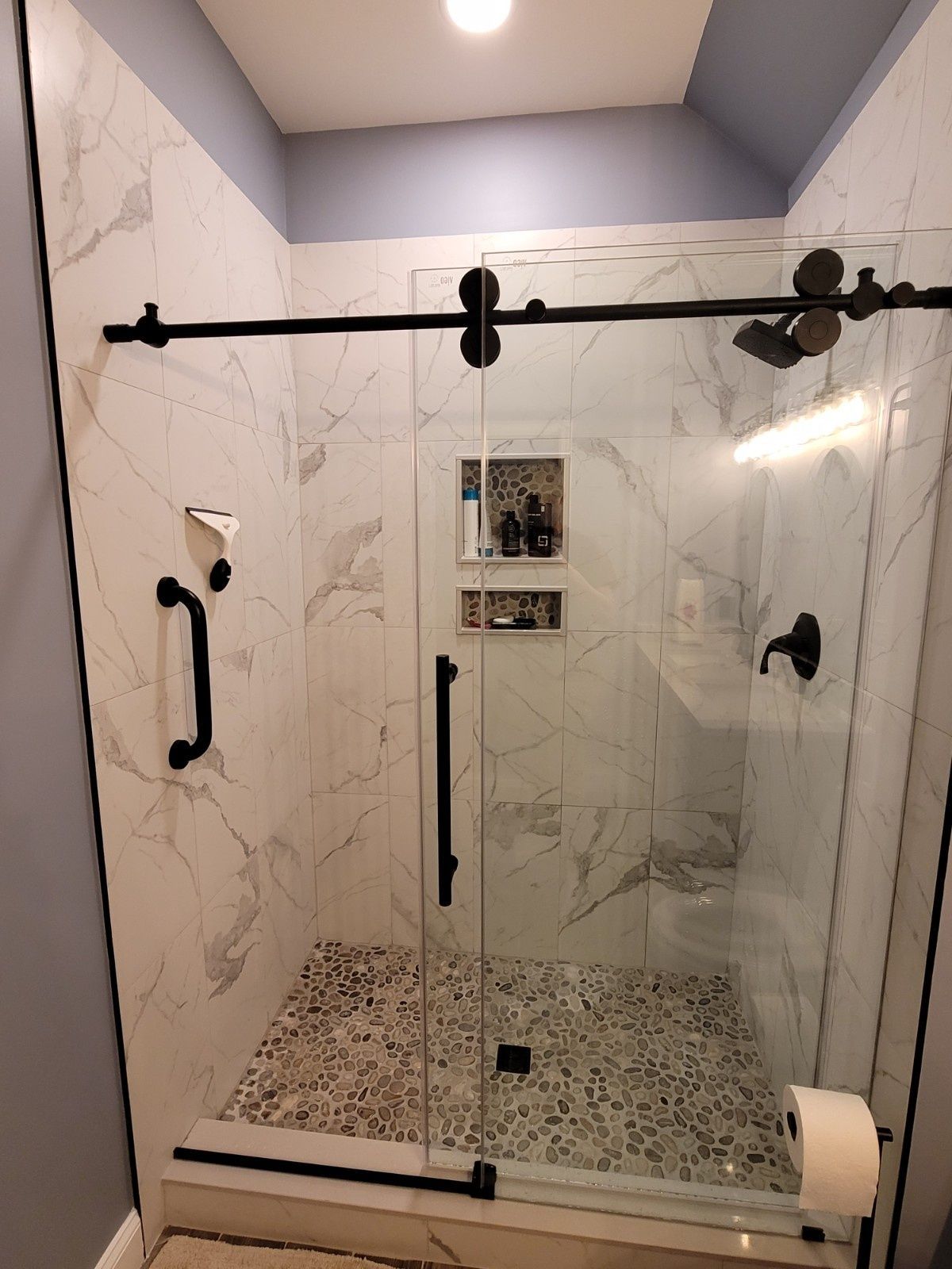 A walk-in shower with white marble-patterned tiles, pebble flooring, black fixtures, a sliding glass door, and grab bar.