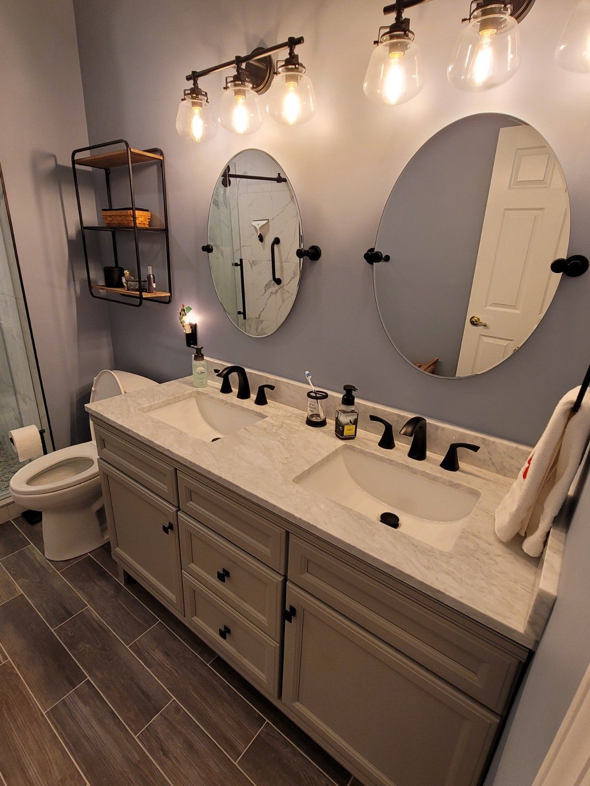 A modern bathroom with a double-sink vanity, light grey cabinets, stone countertop, two oval mirrors, and black fixtures.