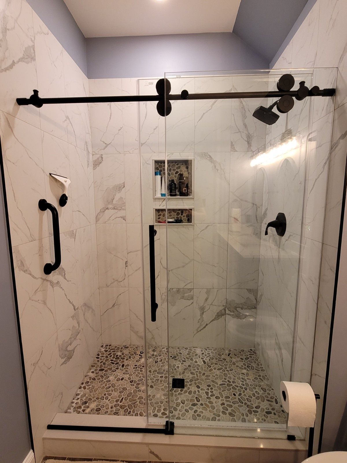 A walk-in shower with marble-look tiles, pebble flooring, a black sliding glass door, and a black grab bar.