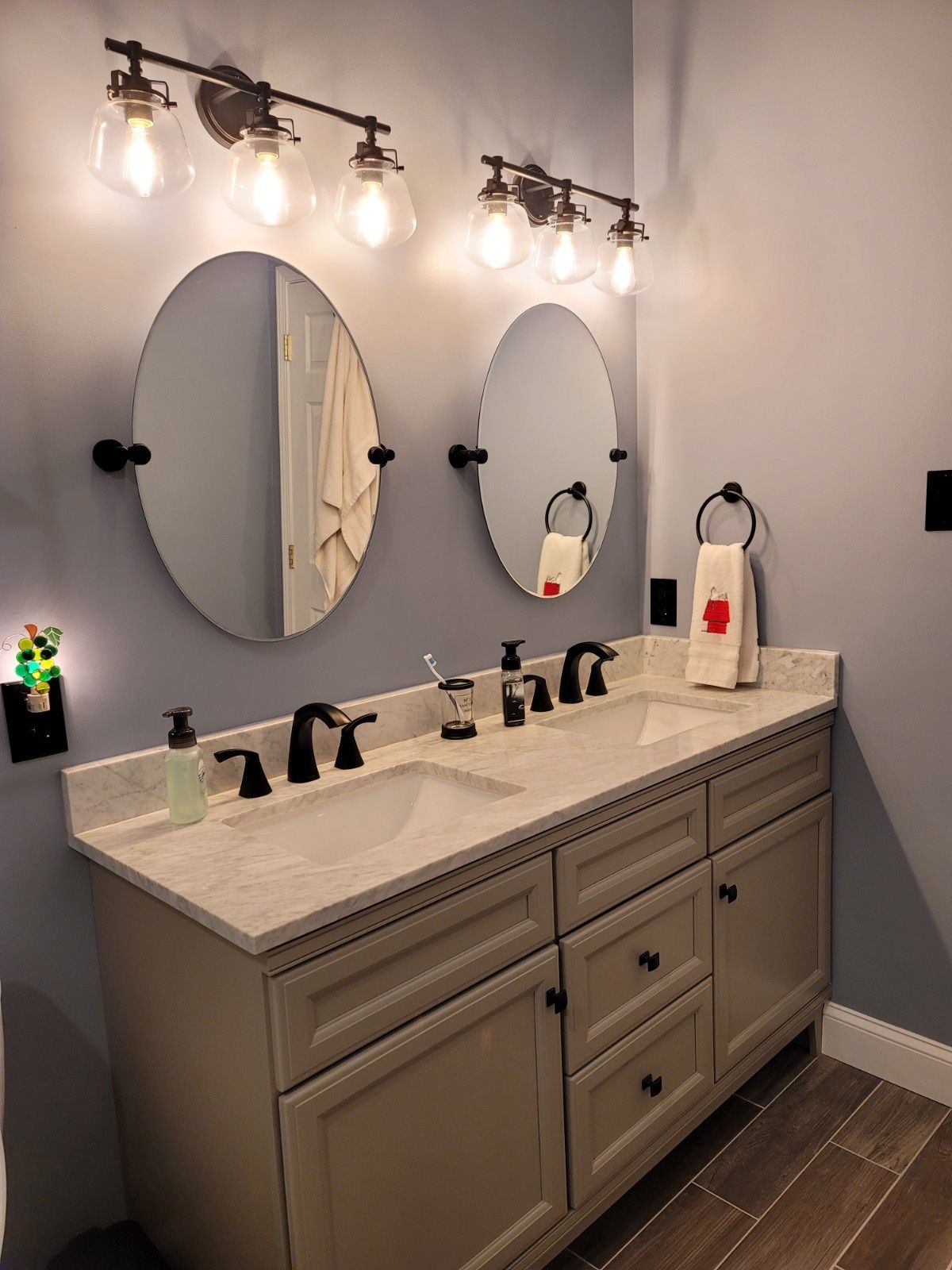 A bathroom vanity with beige cabinets, a light stone countertop, dual sinks, oval mirrors, and black light fixtures.