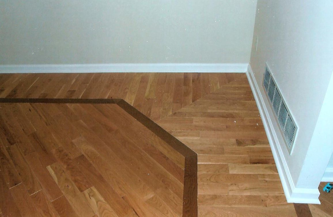 Hardwood floor with a contrasting dark brown border separating two sections of light oak planks and a wall-mounted vent.