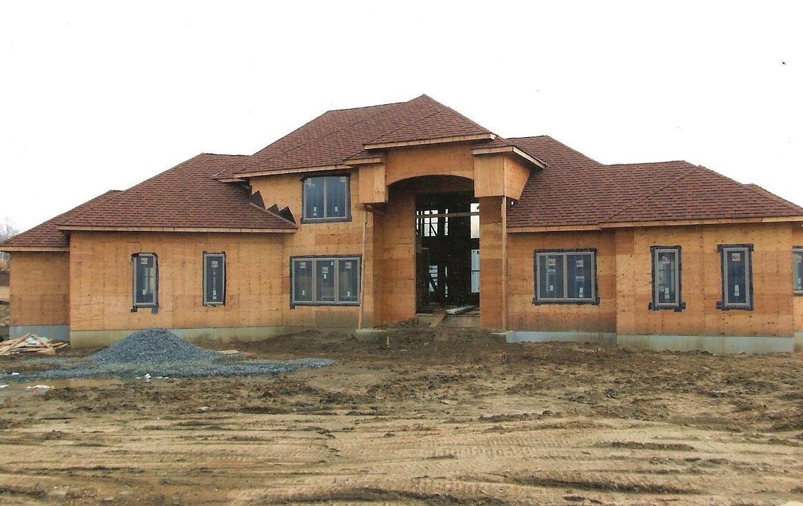 A house under construction with brown-orange walls, a red-tiled roof, and installed windows, set on a dirt plot.