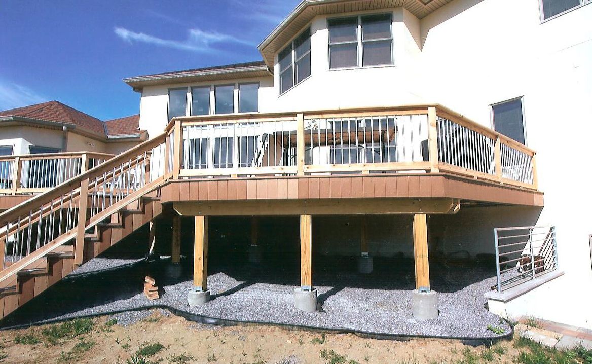 A tan house with a large, elevated wooden deck supported by posts over a gravel yard, with stairs leading up to the side.