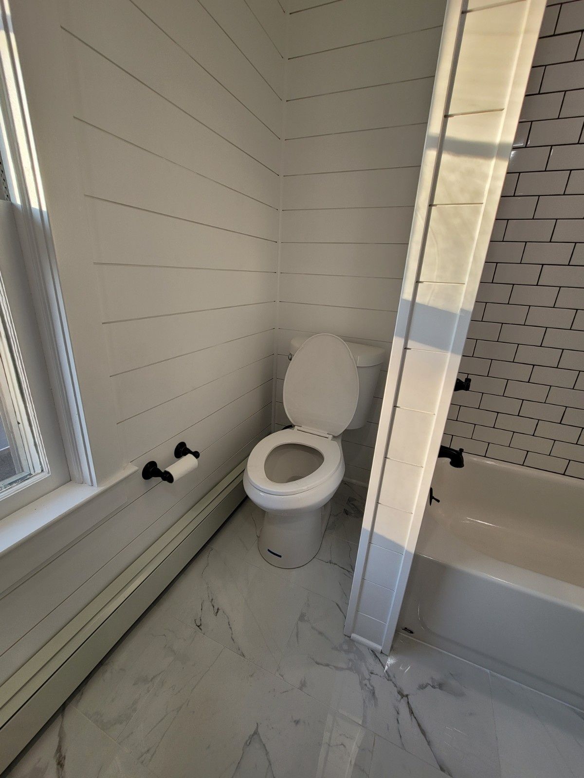 A bathroom with white horizontal plank walls, marble tile floors, a white toilet, and a bathtub with white subway tiles.