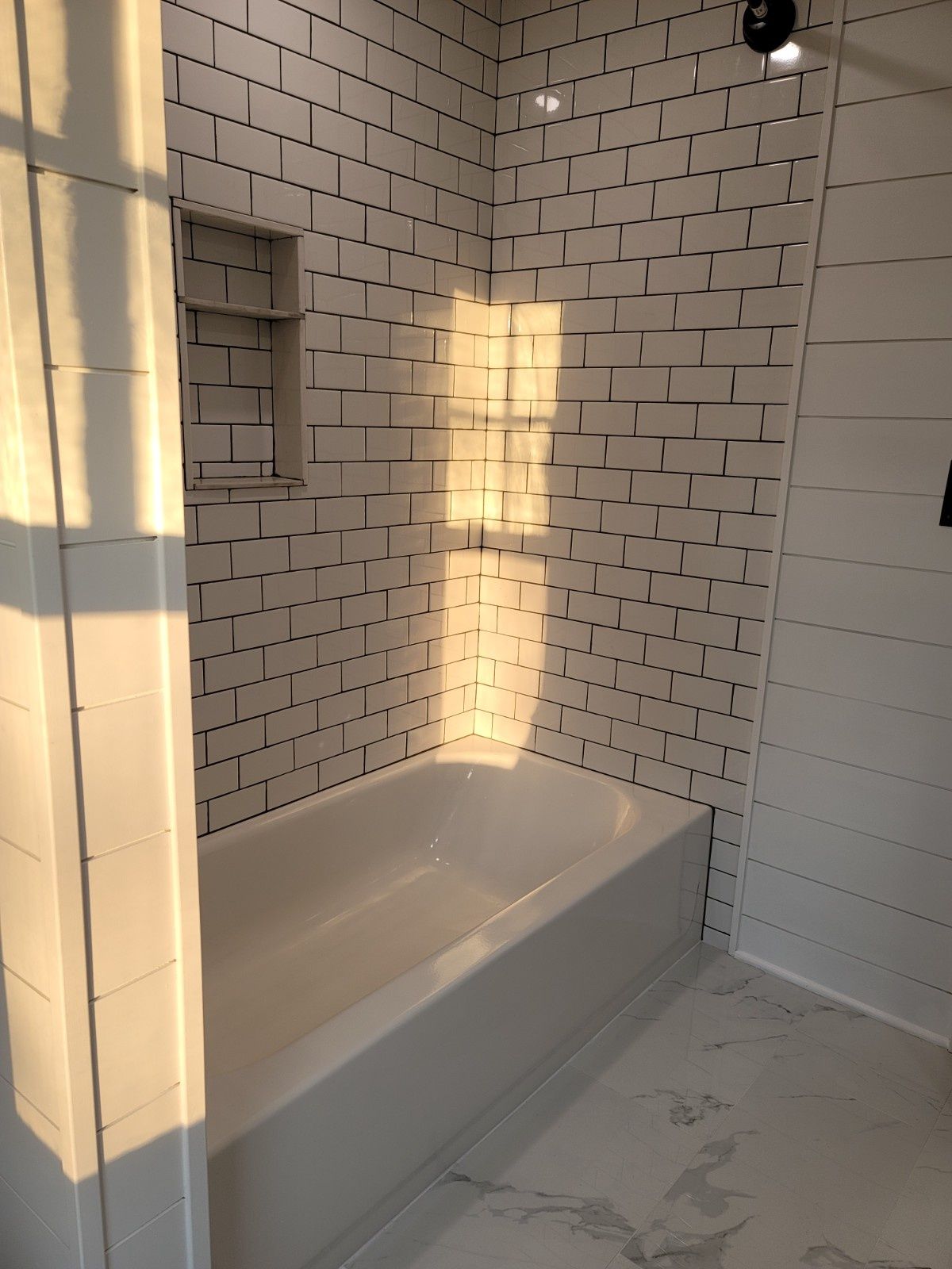 A bathtub set against white subway tile walls with a built-in shelf, bathed in warm, golden sunlight.