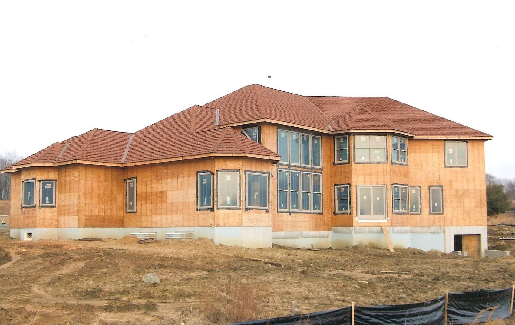A two-story wooden house under construction, featuring a complex tiled roof and multiple windows, set on a dirt lot.