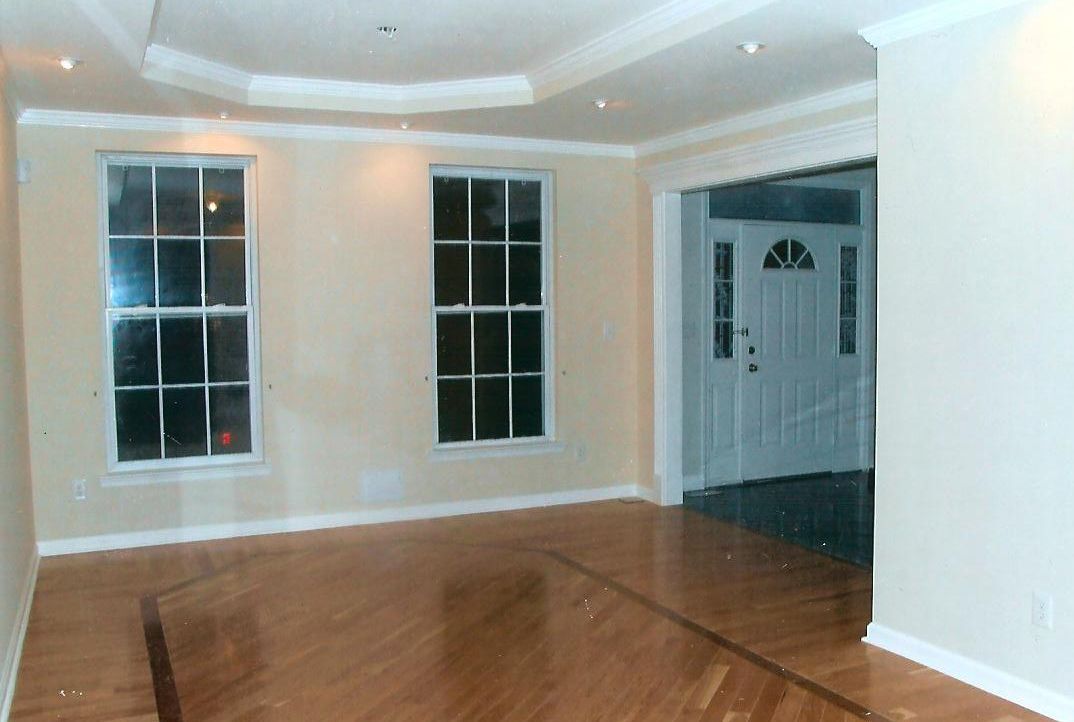 A spacious room with light walls, hardwood floors, a tray ceiling with recessed lights, two windows, and a front entry.