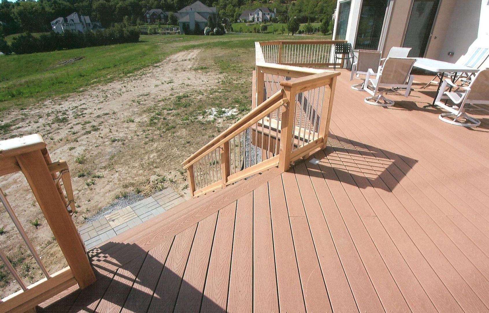 A brown composite deck with wooden railings and stairs leading down to a dirt backyard on a sunny day.