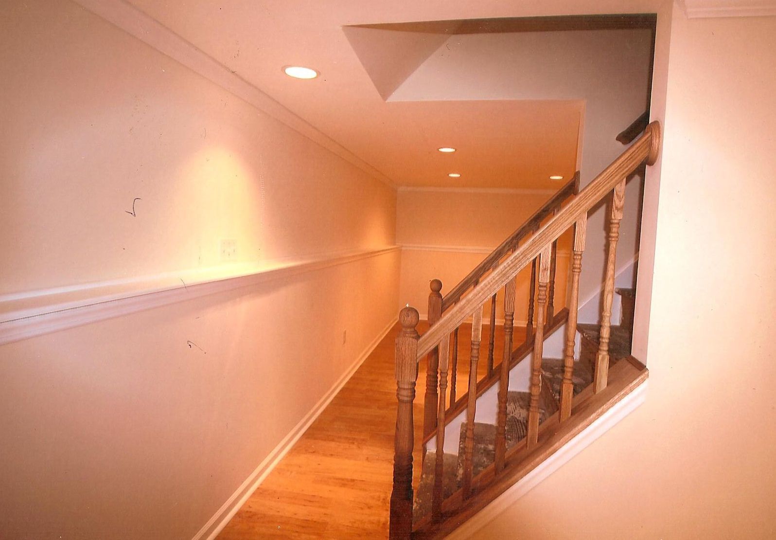 Interior view of a hallway featuring hardwood floors, light-colored walls with chair railing, and a wooden stair railing.