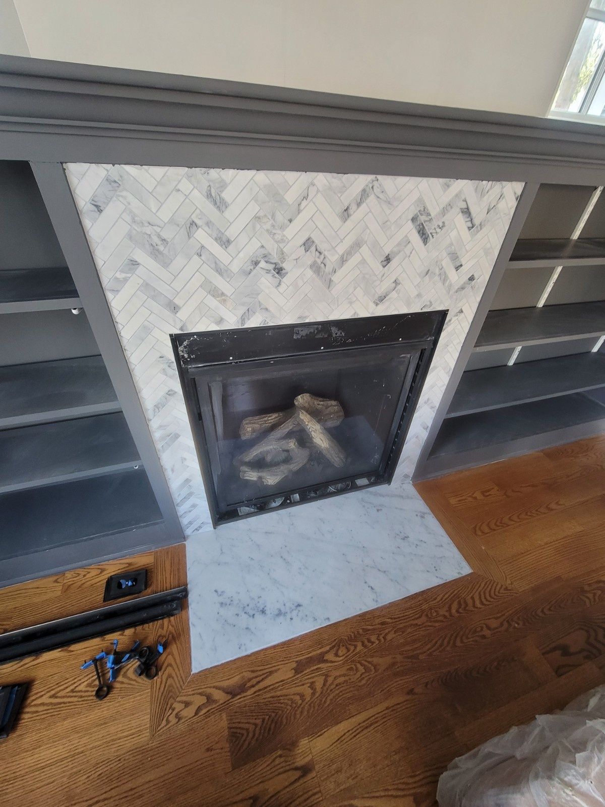 A fireplace featuring a white marble herringbone tile surround and hearth, framed by grey built-in shelving on wood floors.
